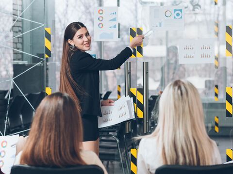 A young businesswoman in a smart black suit is giving a presentation in a modern office setting. She points at various colorful charts and graphs displayed on clear glass panels, explaining data trends to her audience