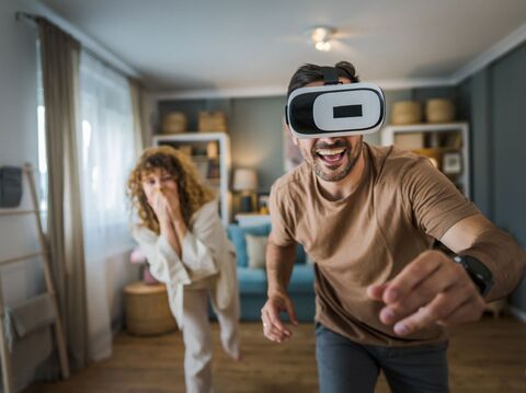 Excited man using a VR headset in living room, playfully engaging with virtual reality as amused woman watches