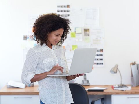 A woman with curly hair using a laptop in a well-lit office, with a whiteboard full of notes in the background