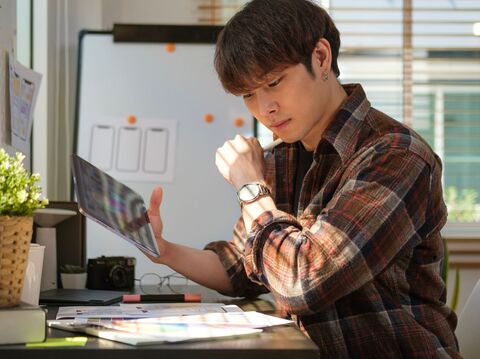 A young man in a plaid shirt working on a tablet with design sketches on a desk