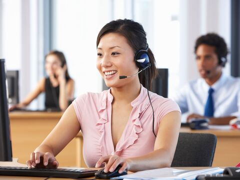 A woman is sitting in the call center, wearing a headset with a microphone, and using a mouse and keyboard. Two people are sitting behind her, which is blurred in image