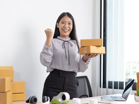 A woman celebrating with a fist pump while holding two boxes, standing in an office with more boxes around