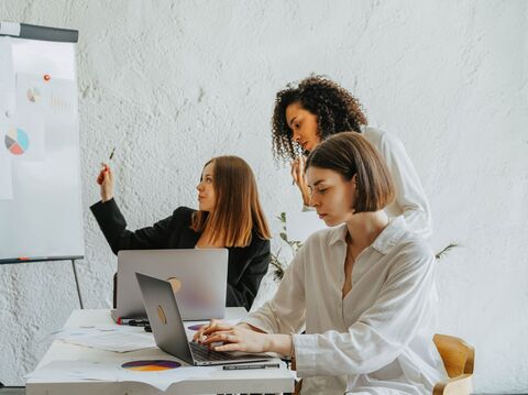 Three women collaborate in an office, one presenting at a flip chart, another typing on a laptop, and the third observing