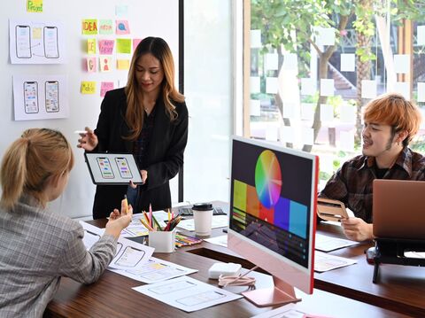 A vibrant team meeting in a creative workspace, featuring a diverse group of three professionals discussing a mobile app design. A woman leading the session, dressed in a black blazer over a plaid shirt, holds a tablet displaying app interfaces to a blonde woman and an orange-haired man seated at the table