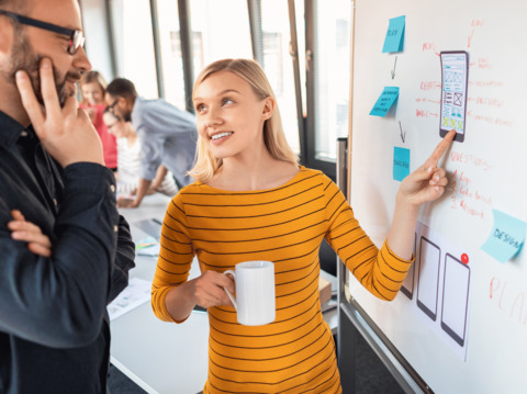 A woman in a yellow striped top explaining a design concept on a whiteboard to a colleague