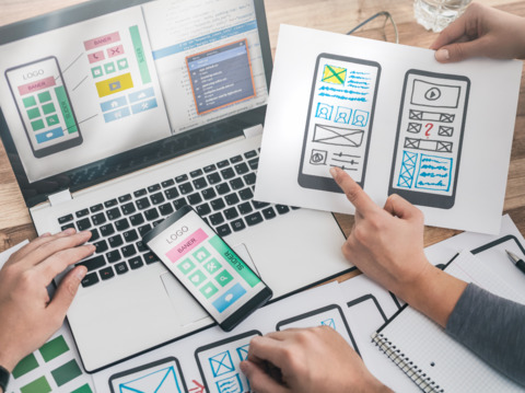 Team designing website or app UI mockups on laptop with wireframes sketched on paper