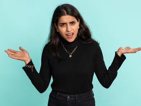 A woman with an inquisitive expression, wearing a black turtleneck and gold necklace, standing against a light blue background, gesturing with both hands