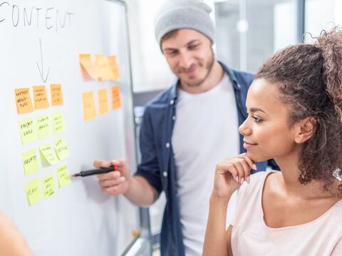 Two people, one holding a marker, brainstorming ideas and concepts on a whiteboard covered with sticky notes