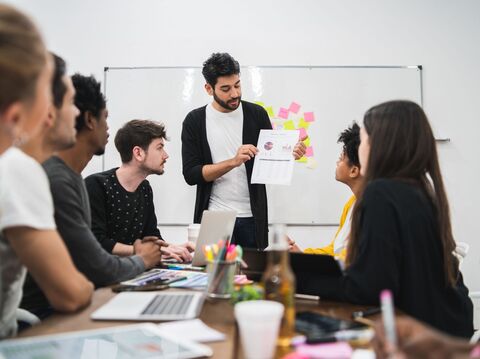 A diverse team of six young professionals gathered around a table in a bright, modern meeting room. A man is standing and presenting a document with charts to his attentive colleagues, who are listening and discussing