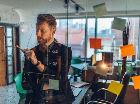 A professional standing in front of a glass wall with post-it notes and diagrams, writing with a marker