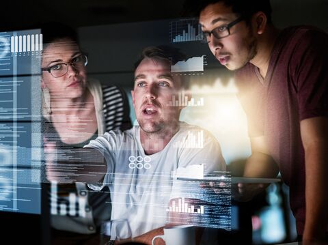 Three coders or developers analyzing data and code visualizations on multiple computer screens in a dimly lit room