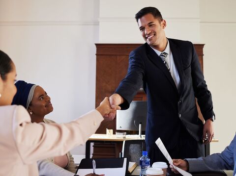 A professional meeting in a boardroom where a man in a dark suit is smiling and shaking hands with a woman wearing a blue headscarf. Other diverse team members are seated around the table, participating in the discussion