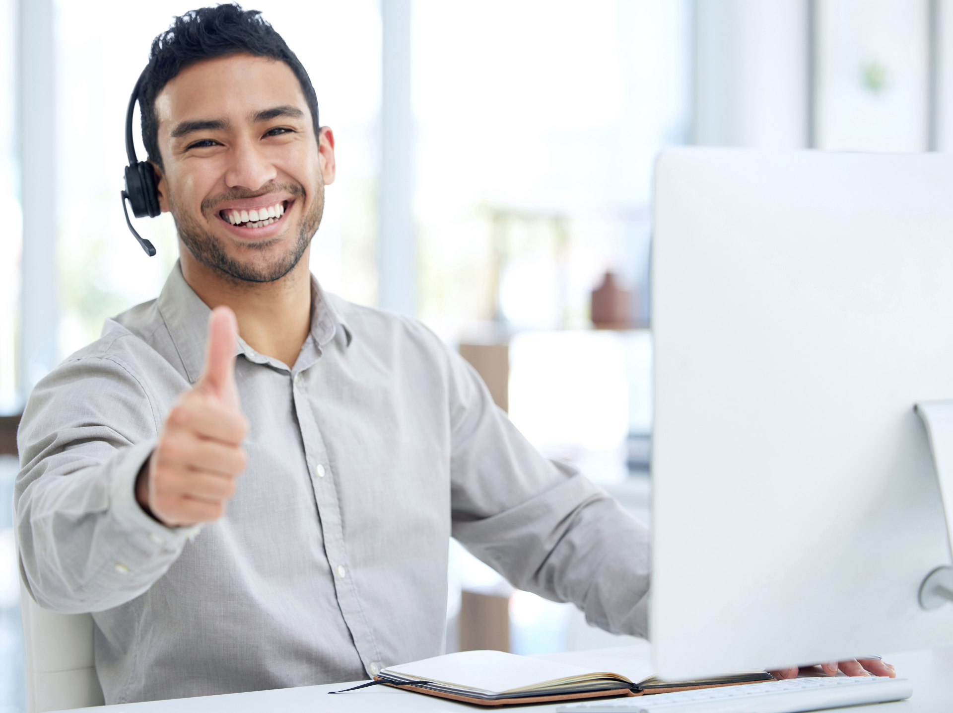 A smiling customer service agent wearing a headset gives a thumbs-up in a bright office setting, conveying positive support and customer satisfaction.
