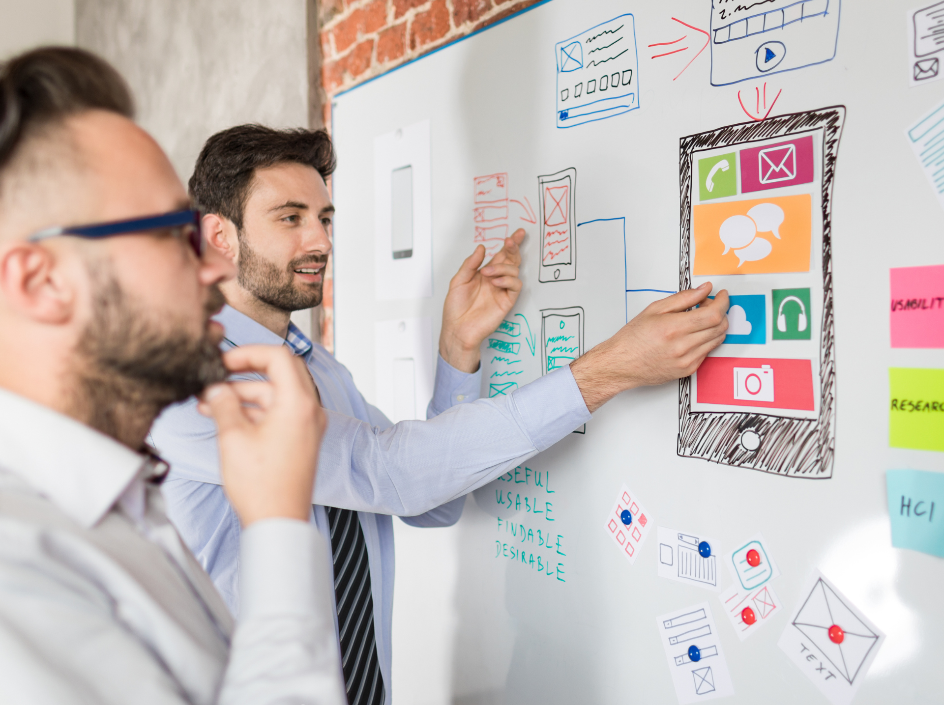 Two men stand in front of a whiteboard, discussing app prototyping on the board's surface