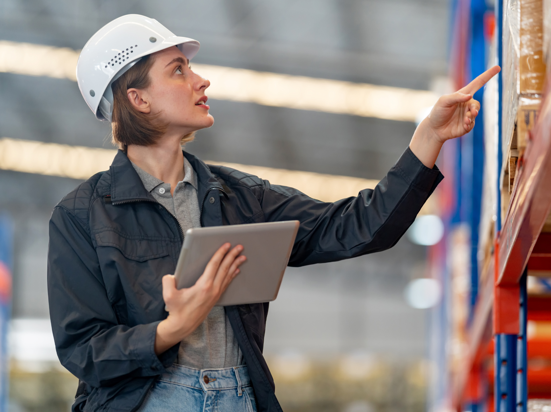 A woman wearing a hard hat and jacket is holding a tablet, focused on her work in a construction or industrial setting