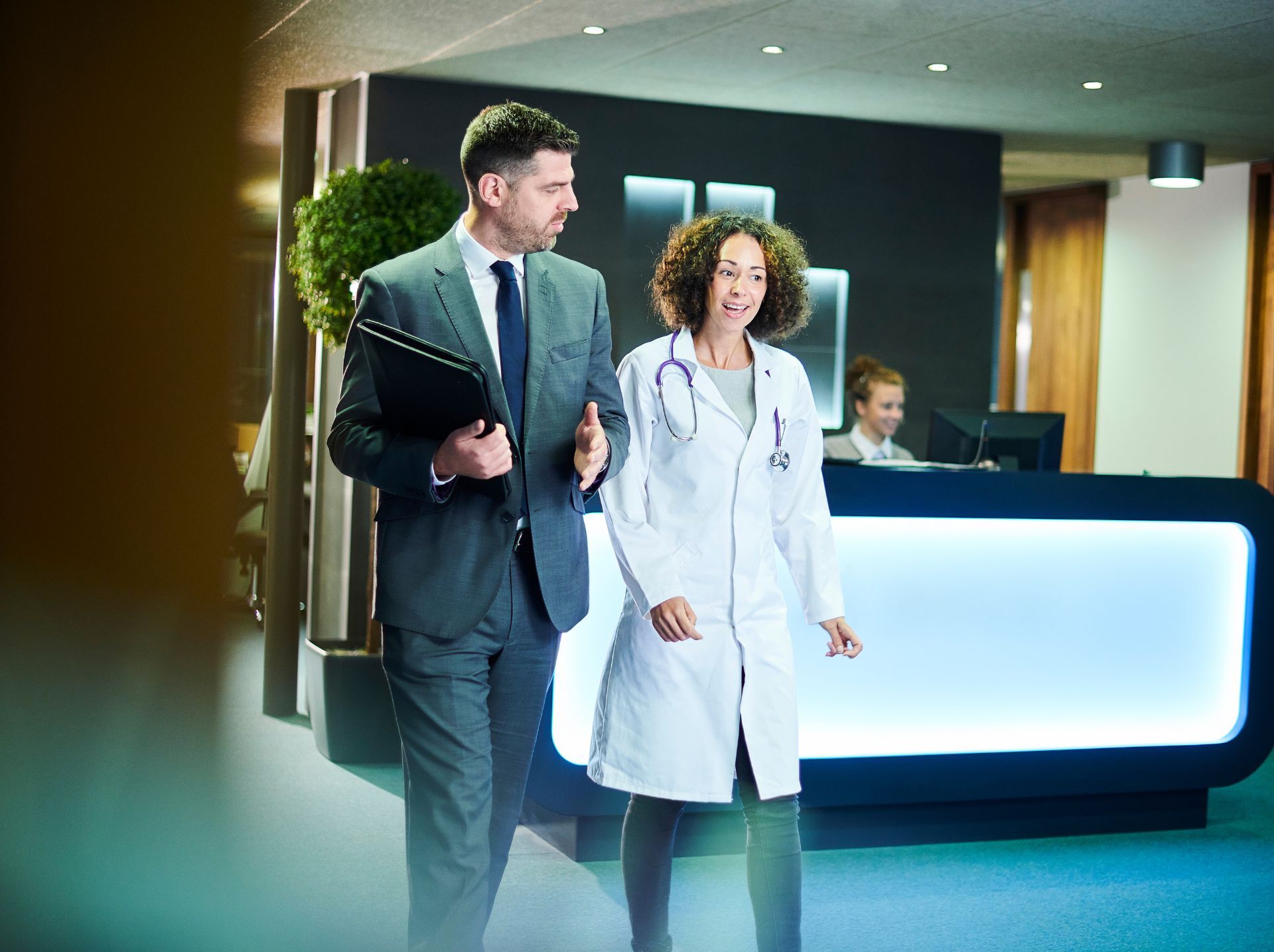 A business professional in a suit is walking alongside a doctor in a white coat. They appear to be having a conversation in a modern medical office reception area with a receptionist working at a desk in the background