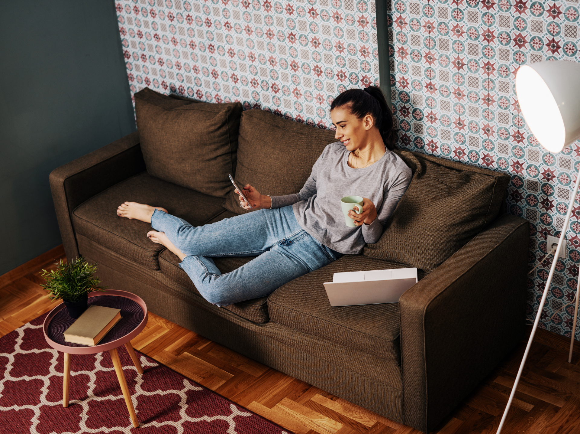 A woman relaxing on a couch with a cup of coffee, browsing her smartphone, with a laptop beside her in a cozy living room setting