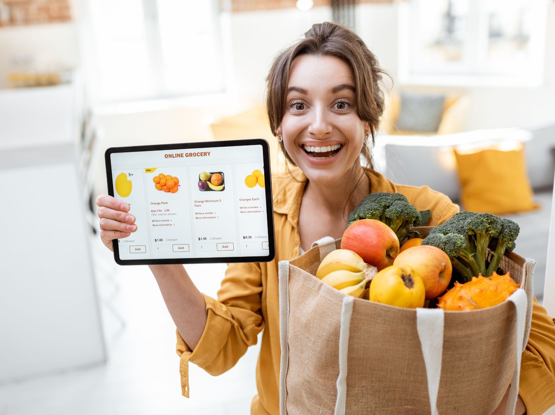 A woman is smiling and holding a tablet showing an online grocery shopping interface with various fruits listed. In her other hand, she is holding a bag filled with fresh fruits and vegetables. The background appears to be a bright, modern kitchen