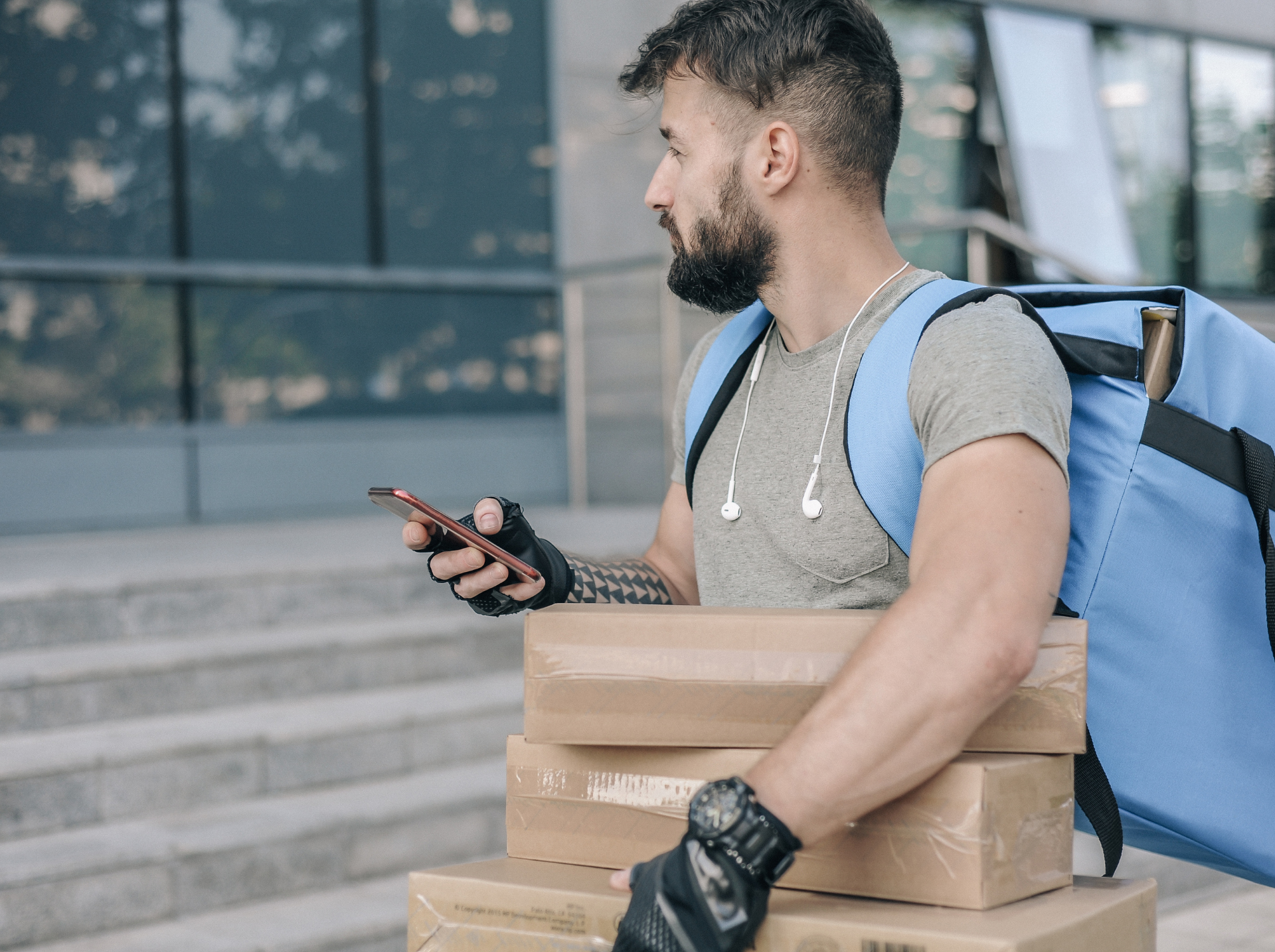 A person with a beard holds several packages, wearing a blue backpack and headphones, looking at a smartphone.