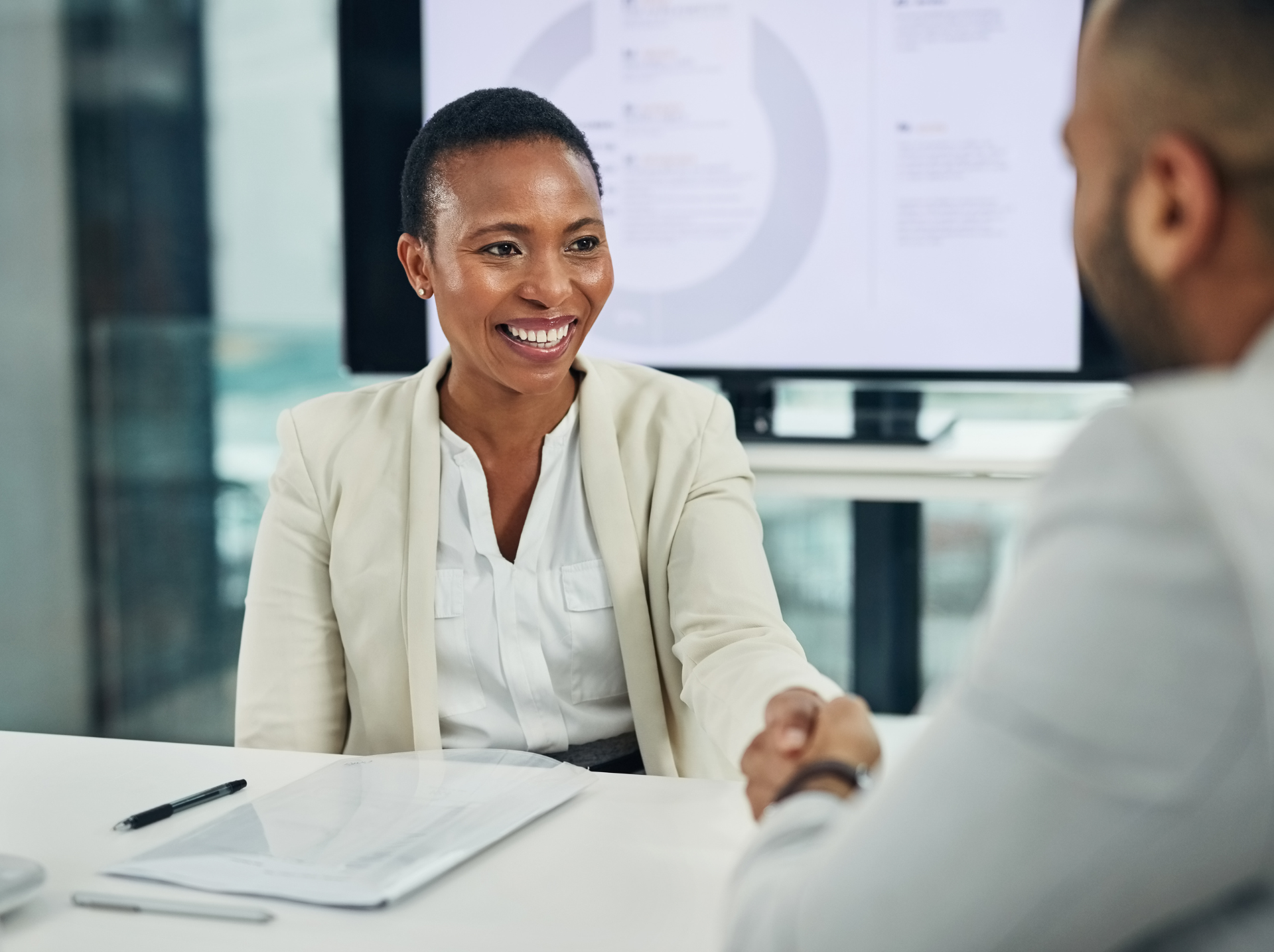 A professional woman in business attire smiles and shakes hands with a colleague across a desk, symbolizing a successful business agreement or collaboration.