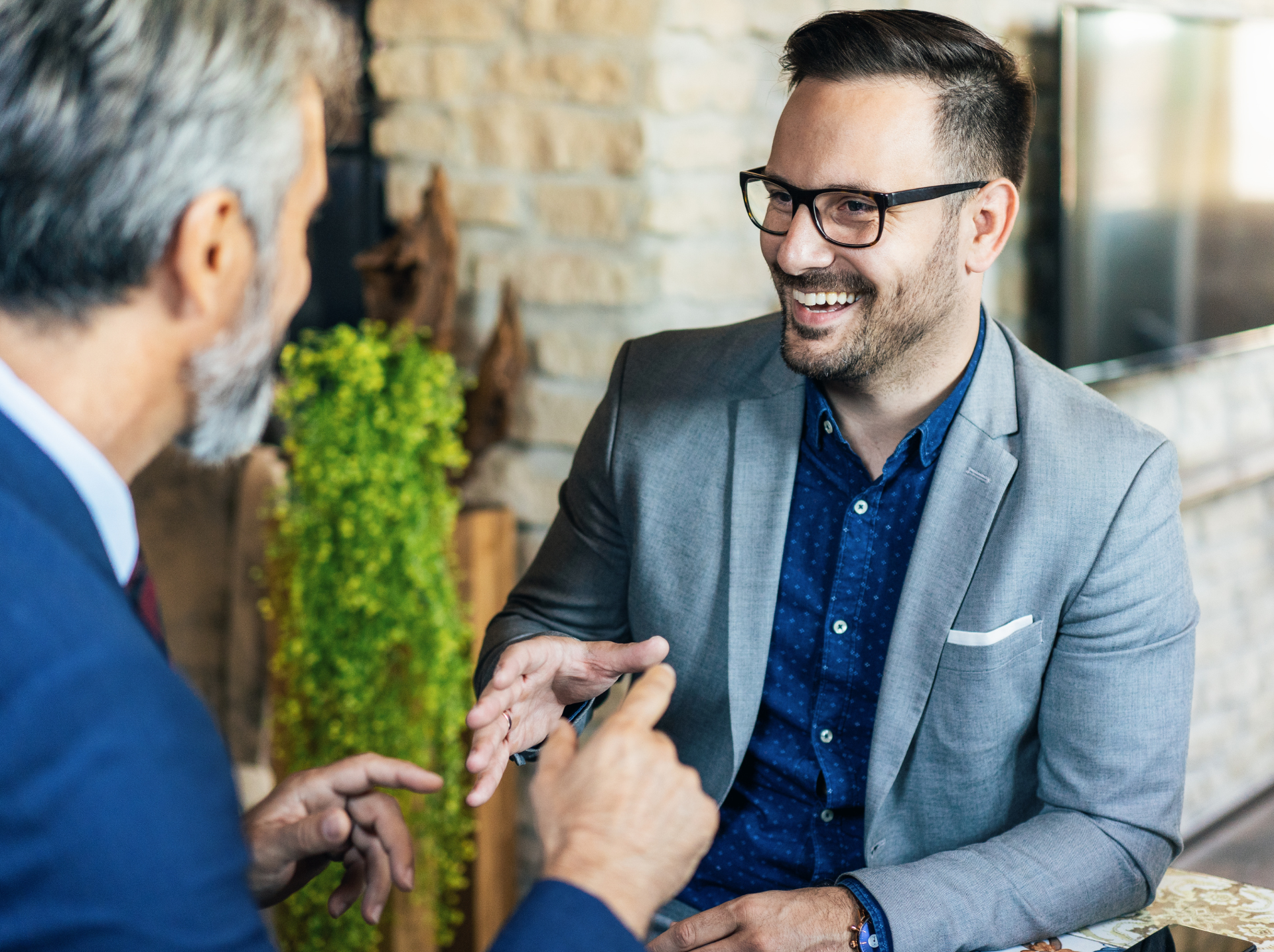 Two men in business attire are engaged in a lively conversation. One man is smiling, wearing glasses and a gray suit, while the other gestures with his hand. They are indoors with brick walls.